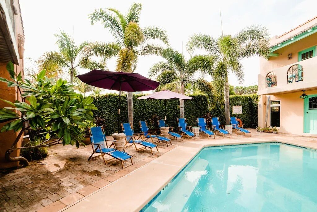 A pool area with blue lounge chairs and umbrellas, surrounded by palm trees and a building with a green door.