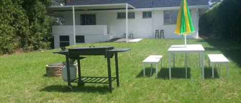 A grassy outdoor area with a barbecue table, a gas cylinder, and a green and yellow umbrella.