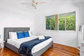 Ein Schlafzimmer mit Bett, Deckenventilator, Fenster mit Blick auf Grünflächen und einer weißen Wand.