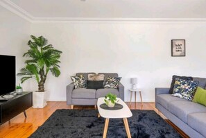Ein modernes Wohnzimmer mit einem grauen Sofa, einem schwarzen Teppich, einem weißen Couchtisch, einer großen Topfpflanze und einem gerahmten Zitat an der Wand.