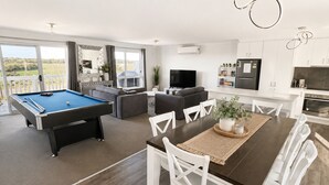 Un salon moderne avec un billard, un coin repas avec des chaises blanches, une cuisine avec des meubles blancs et une grande fenêtre donnant sur un paysage verdoyant.