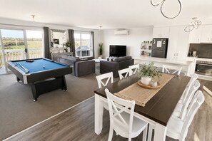 A modern living room with a pool table, a dining area with white chairs, a kitchen with white cabinets, and a large window with a view of greenery.