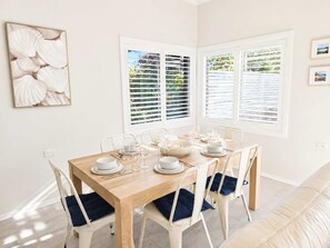 Un comedor con una mesa de madera dispuesta para cuatro personas, sillas blancas con cojines azules y una obra de arte enmarcada sobre la mesa.