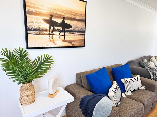 Un salon avec un tableau encadré représentant des surfeurs sur une plage, un canapé gris agrémenté de coussins bleus et à motifs, une table d’appoint blanche avec une plante et un objet blanc, et un panier en osier.