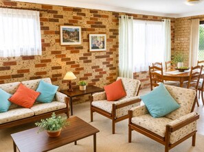 Un salón con pared de ladrillo, dos sofás con cojines estampados, una mesa de centro de madera y una zona de comedor con mesa y sillas.
