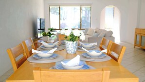 A dining table set with plates, bowls, and a vase of flowers, surrounded by chairs in a bright room with a sofa and a TV in the background. - Sandy Shore Villa at Mollymook (Mollymook)