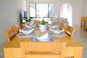 A dining table set with plates, bowls, and a vase of flowers, surrounded by chairs in a bright room with a sofa and a TV in the background.