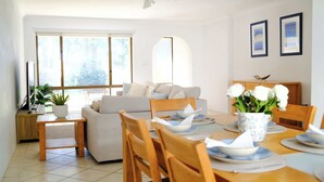 A modern living room with a sofa, dining table, and chairs, a potted plant, and a window with a view. - Sandy Shore Villa at Mollymook (Mollymook)
