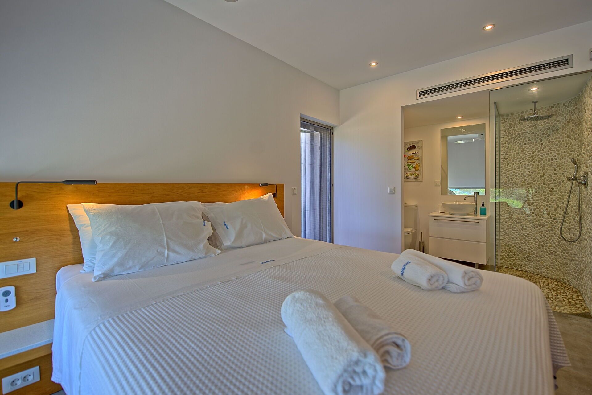 Quarto de hotel com uma cama grande, cabeceira de madeira, banheiro com box de vidro e pia branca.
