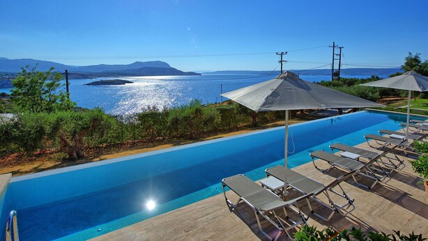 Ein Pool mit Liegestühlen und Sonnenschirmen, mit Blick auf ein Gewässer und Berge in der Ferne.