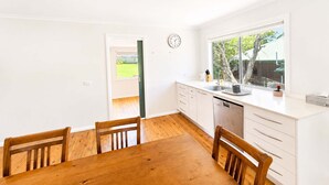 A bright kitchen with a wooden dining table and chairs, a window with a view of greenery, and a clock on the wall. - Harbour Haven at Ulladulla (Ulladulla)