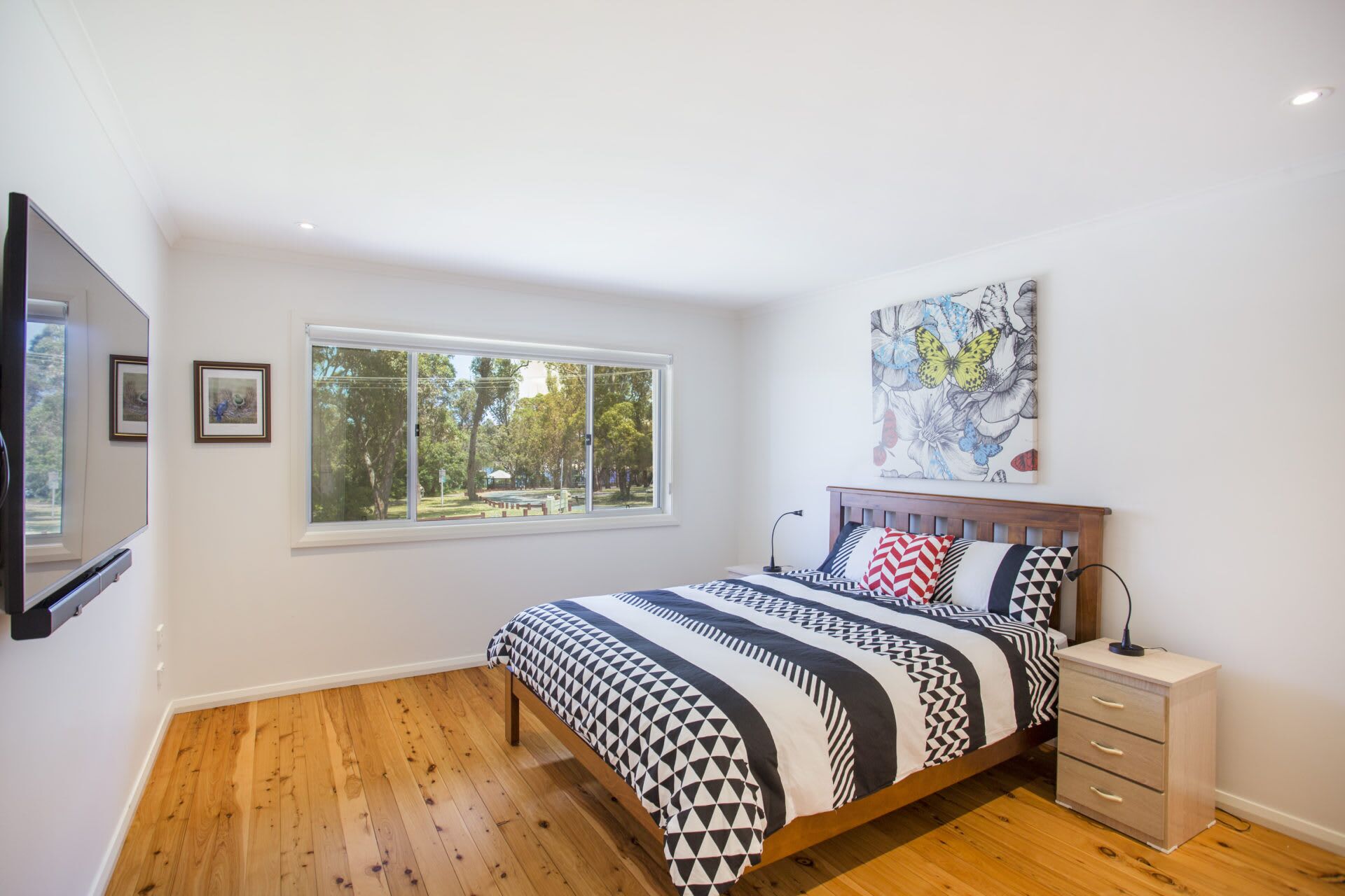 A bedroom with a bed, a nightstand, a wall-mounted TV, a window with a view of trees, and a painting on the wall.