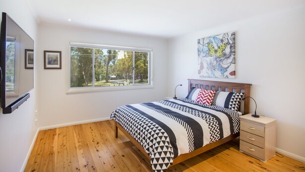 A bedroom with a bed, a nightstand, a wall-mounted TV, a window with a view of trees, and a painting on the wall. - Harbour Haven at Ulladulla (Ulladulla)