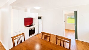 A compact kitchen with white cabinets, a red backsplash, and a dining area with a wooden table and chairs. - Harbour Haven at Ulladulla (Ulladulla)