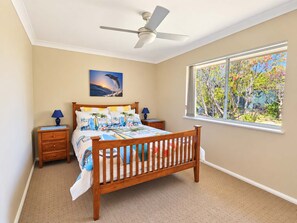 A bedroom with a wooden bed, a ceiling fan, a window with a view of trees, and a painting on the wall.
