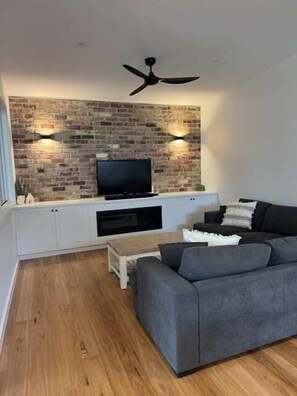 A modern living room with a brick accent wall, a flat-screen TV, a grey sofa, and a ceiling fan.