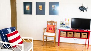 Un salon avec un canapé blanc, une chaise en bois avec un coussin coloré, un meuble TV rouge avec des paniers, et trois tableaux encadrés accrochés au mur.