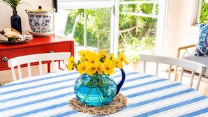 Une table à manger recouverte d’une nappe à rayures bleues et blanches, un vase bleu contenant des fleurs jaunes, et un buffet rouge en arrière-plan.