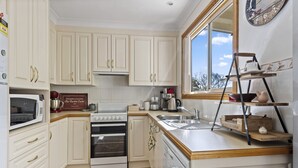 A kitchen with white cabinets, a stove, a microwave, a sink, and a window with a view. - Jillbys House at Narrawallee (Narrawallee)