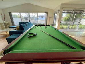 Una mesa de billar en una habitación con un sofá azul, una mesa de centro de vidrio y ventanas amplias.
