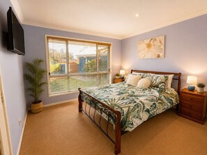 Un dormitorio con una cama, una ventana con persianas, un televisor montado en la pared, una mesita de noche con lámpara y una planta en maceta.
