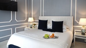 Un lit bien fait, recouvert d’une couette blanche et marine, deux tables de chevet avec des lampes, un téléviseur à écran plat fixé au mur et un plateau avec des fruits et des viennoiseries posé sur le lit.