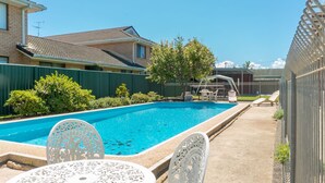 Ein rechteckiger Swimmingpool mit klarem, blauem Wasser, umgeben von einer Betonplattform. Zwei weiße Tische und Stühle stehen draußen, und es gibt eine überdachte Sitzgelegenheit mit Tisch und Stühlen.
