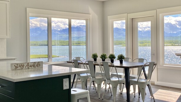 A dining area with a view of a lake and mountains, white chairs, a wooden table, and a sign that reads "stay Awake". - Brand new beautiful home with best view outside of Waterton Park. UPPER suite. (Mountain View)