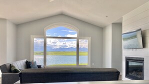 A modern living room with a large window offering a view of a lake and mountains. - Brand new beautiful home with best view outside of Waterton Park. UPPER suite. (Mountain View)