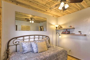 A bedroom with a wooden ceiling, a bed with patterned bedding, a ceiling fan, a wall-mounted mirror, and a shelf with books and a plant. - Tasteful Log Cabin Retreat on Lake Pagosa! (Pagosa Springs)