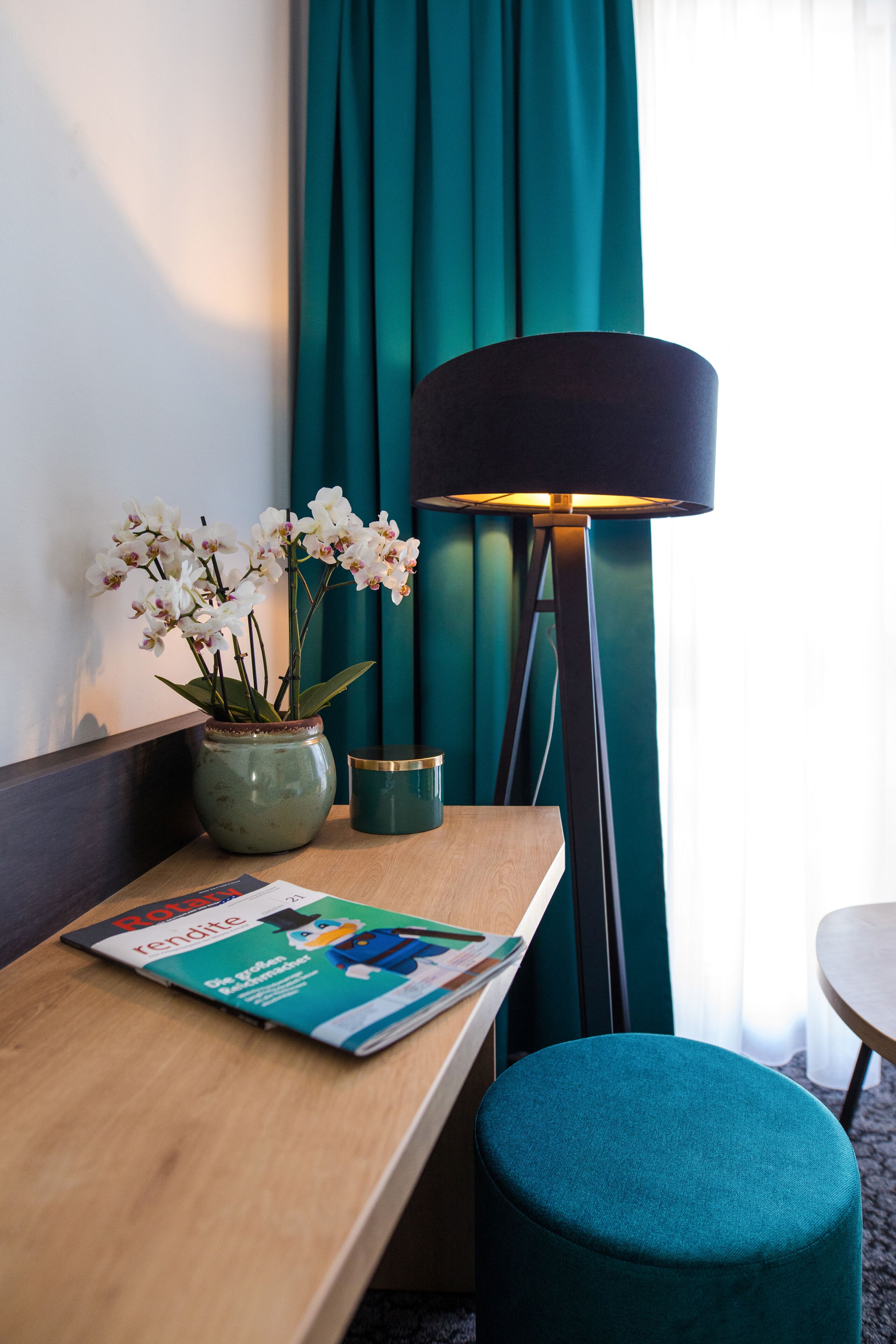 Habitación de hotel con un escritorio de madera, un reposapiés color turquesa, un jarrón con flores blancas, una lámpara y cortinas color turquesa.