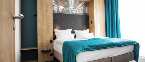 A modern hotel room with a bed, wooden paneling, a large window with curtains, and a view of an industrial area.