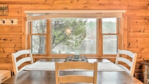 L’intérieur d’un chalet en bois comprend une table à manger, deux chaises et une vue sur un lac, visible à travers de grandes fenêtres.