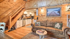 L’intérieur chaleureux d’un chalet avec un escalier en bois, un coin salon doté d’un canapé et de coussins, une cuisine et un grand tableau encadré accroché au mur.