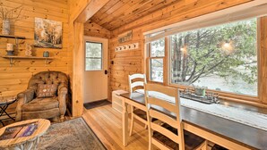 L'intérieur chaleureux d'un chalet, avec un revêtement mural en bois, une table à manger entourée de chaises, une cheminée et une grande fenêtre donnant sur la forêt.