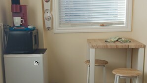 Petita cuina compacta amb nevera, microones i cafetera. Hi ha una taula petita amb dos tamborets i una finestra amb persianes.