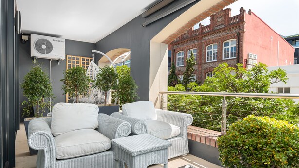 A balcony with wicker furniture, a potted plant, and a view of a red brick building. - Rockwell 114 (Cape Town)