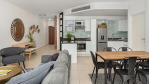 A modern living room with a grey sofa, a wooden dining table, and black chairs. A kitchen is visible in the background with white cabinets and a microwave. - Rockwell 114 (Cape Town)
