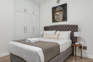 A neatly made bed with a grey headboard, white bedding, and a brown throw blanket. There is a small bedside table with a lamp and a wall-mounted artwork above the bed. - Rockwell 114 (Cape Town)