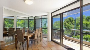 Enjoy meals at the large dining table surrounded by wicker chairs, with a view of lush greenery outside through expansive windows.