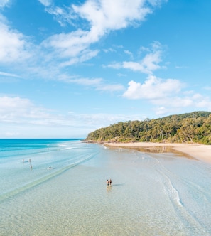 Una spiaggia con acqua cristallina, persone che passeggiano e una collina ricoperta di alberi.