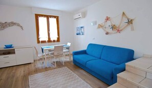 Una habitación con un sofá azul, un escritorio con una computadora portátil, una silla blanca y una mesita con un tazón azul.