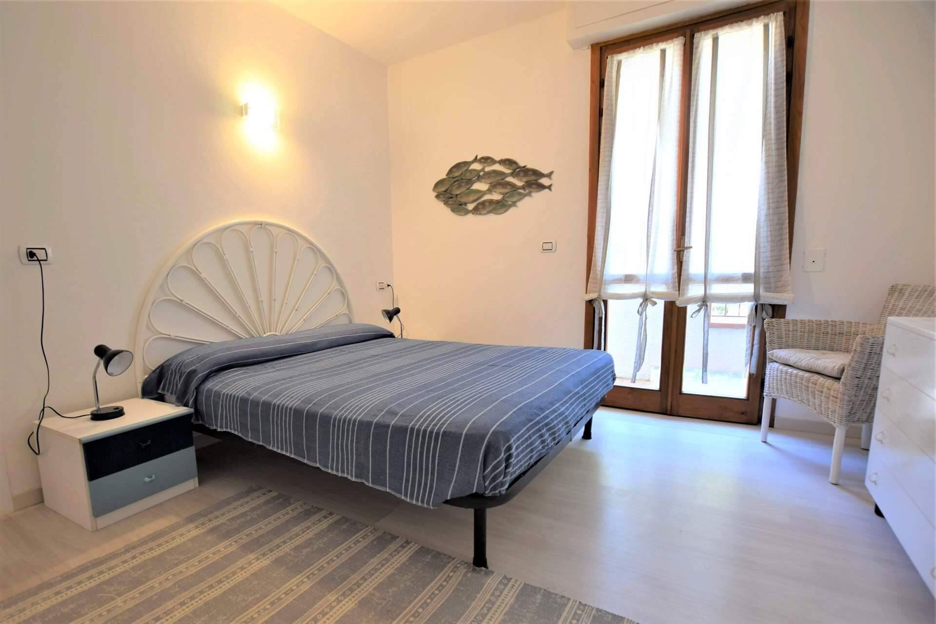 Ein Schlafzimmer mit Bett, Nachttischen, einer Lampe, Wanddeko, einem Fenster mit Vorhängen und einem Sessel.
