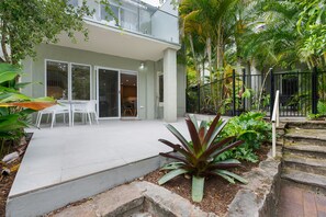 Una casa moderna a due piani con un patio, sedie bianche e diverse piante tropicali.