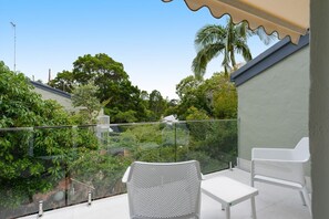 Un balcon agrémenté de chaises et d’une table en osier blanc, entouré de rampes en verre et d’une végétation luxuriante.