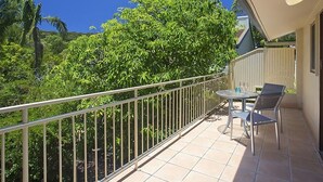 A balcony with a metal railing, a small table with chairs, and a view of lush greenery and a clear blue sky.