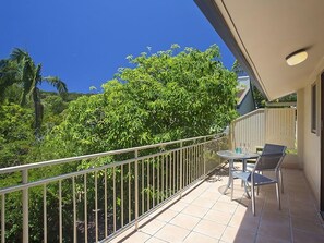 Un balcone con ringhiera metallica, un tavolino con sedie e vista su una vegetazione lussureggiante e un cielo azzurro intenso.