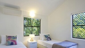 A bedroom with two beds, a nightstand, a lamp, and windows with a view of greenery.