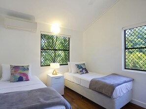 A bedroom with two beds, a nightstand, a lamp, and windows with a view of greenery.