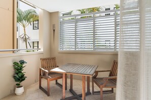 Uma varanda com mesa e cadeiras de madeira, um vaso de planta e uma janela com persianas.